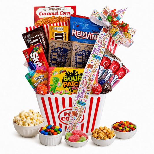A candy and snack gift basket arranged in a red-and-white popcorn bucket with a large decorative bow, surrounded by bowls of popcorn, M&M's, Skittles, Sour Patch Kids, Red Vines, and other assorted sweets on a white background.