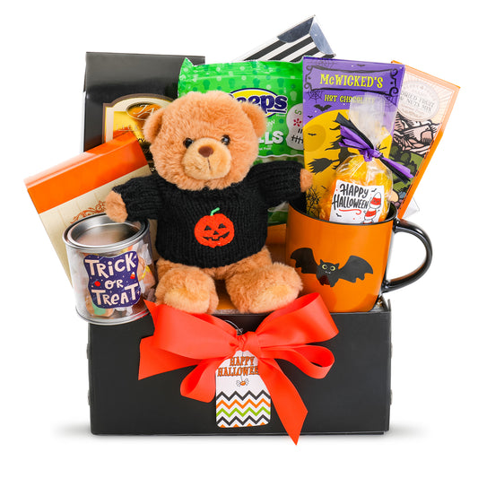 A Halloween-themed gift basket featuring a plush teddy bear in a black sweater with a pumpkin face, an orange mug with a bat design, a tin of trick or treat, packaged snacks, and a black box tied with a bright orange ribbon and a 'Happy Halloween' tag.