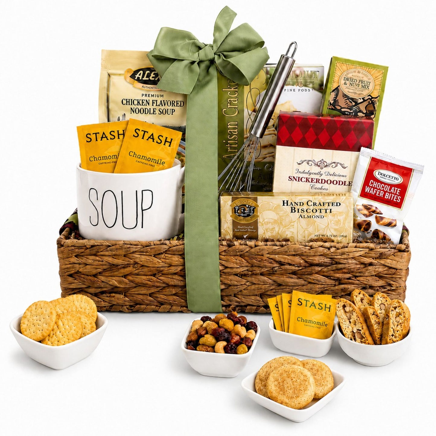 A gourmet gift basket filled with snacks and treats, tied with a large green ribbon. The basket contains a white ceramic mug labeled 'SOUP' with Stash chamomile tea packets, chicken noodle soup, handcrafted biscotti, chocolate wafer bites, dried fruit and nut mix, and other packaged snacks.