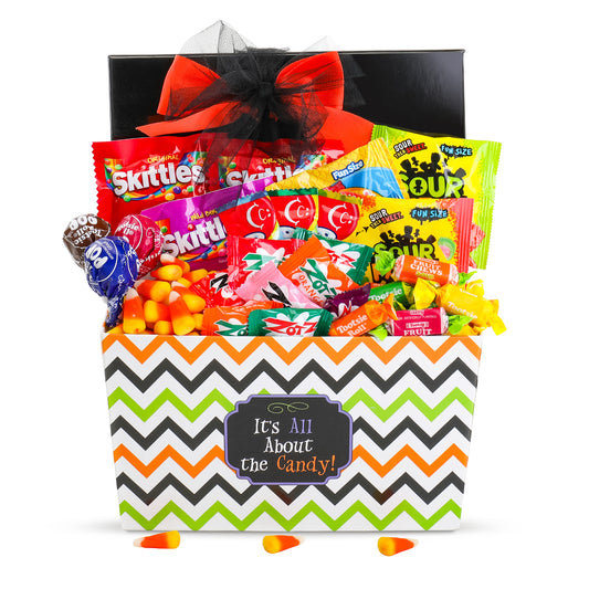 A Halloween-themed candy gift basket featuring a black and orange chevron-patterned box with a black bow and a chalkboard-style label that reads 'It's All About the Candy!'. The basket is filled with assorted colorful candies set against a plain white background.