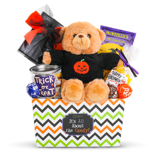 Halloween-themed candy and plush gift basket set featuring a brown teddy bear in a black knit sweater with a pumpkin face, sitting in a white basket with a black, orange, and green chevron pattern. The basket is filled with assorted candies, including McWicked's Hot Chocolate, a can of Trick or Treat Taffy with a black tulle bow.